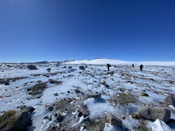 Rocas esparcidas por un campo de hielo, con los científicos buscando meteoritos al fondo